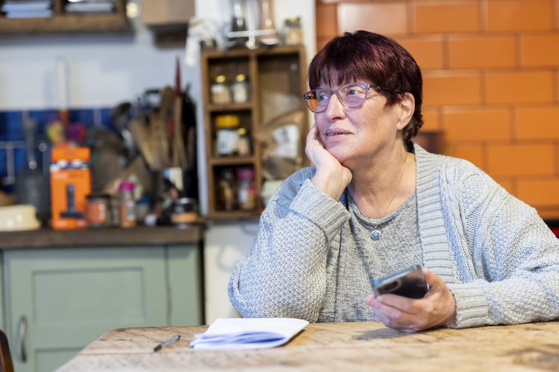 An older woman sits at her kitchen table holding her phone, a stack of paperwork in front of her